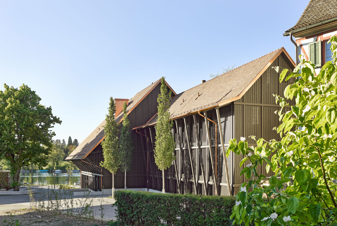 Exterior of Hotel 1802+, a modern barn-style building with dark timber walls and a pitched roof, set in a rural landscape. Guests walk toward the entrance, with an outdoor terrace and trees nearby.