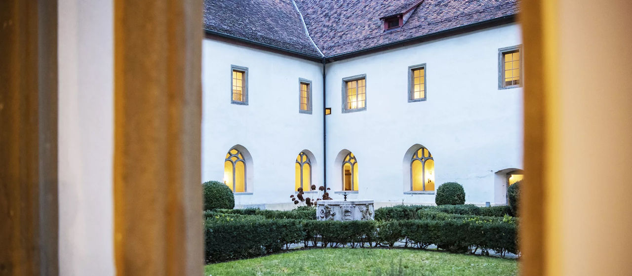 View into the inner courtyard of Kostergut Paradies, showing a historic white building with arched windows glowing with warm light. A small stone fountain sits amid trimmed hedges and greenery, creating a calm, elegant atmosphere.
