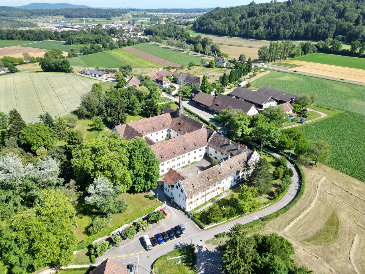 View into the inner courtyard of Kostergut Paradies, showing a historic white building with arched windows glowing with warm light. A small stone fountain sits amid trimmed hedges and greenery, creating a calm, elegant atmosphere.