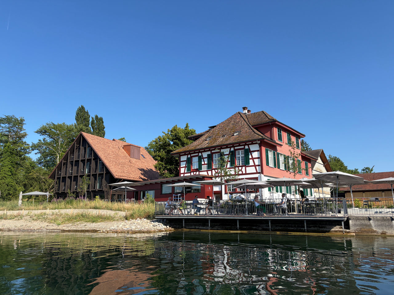 Exterior of Restaurant 1802+, featuring a historic half-timbered building beside a modern extension. The restaurant is set in a green countryside setting with trees, outdoor seating, and warm sunlight.