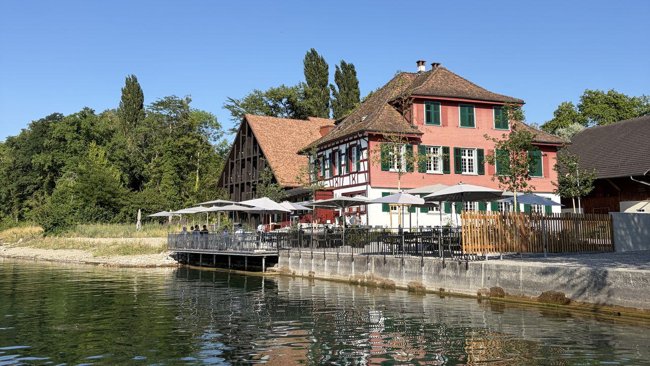 Exterior of Restaurant 1802+, featuring a historic half-timbered building beside a modern extension. The restaurant is set in a green countryside setting with trees, outdoor seating, and warm sunlight.